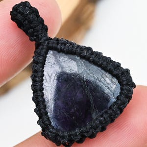 Op de afbeelding: Een driehoekige, donkerpaarse edelsteen hanger, gezet in een zwarte macrame setting. De hanger wordt door een persoon vastgehouden. De tekst "MAYANK JEWELRY" is zichtbaar bovenaan de afbeelding.