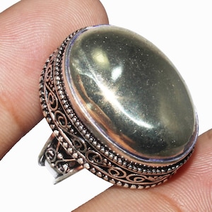Puede incluir: Un anillo de plata con una piedra preciosa de pirita de forma ovalada engastada en un engaste decorativo. El anillo se lleva en un dedo.
