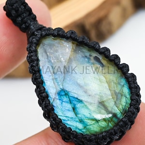 Peut inclure: Un pendentif en forme de goutte d'eau avec une pierre précieuse irisée polie aux teintes bleues et vertes, sertie dans un macramé noir. Le pendentif est tenu dans une main, avec le texte "MAYANK JEWELRY" visible.