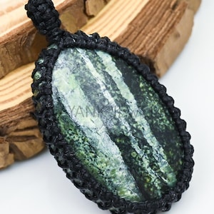 May include: An oval-shaped pendant with a green and white stone, encased in a black woven cord. The stone has a striped pattern. The pendant is displayed on a wooden surface. The text "MAYANK JEWELRY" is visible at the top.