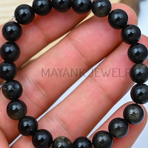 May include: Black beaded bracelet with gold flecks. The bracelet is made of round beads and is strung on a silver cord.