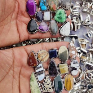 May include: A collection of gemstone rings with silver-colored settings. The rings feature various stone shapes and colors, including purple, blue, green, and brown. The rings are displayed in a hand, with many more rings visible in the background.
