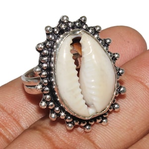 May include: A silver ring featuring a white cowrie shell set within a decorative, beaded frame.