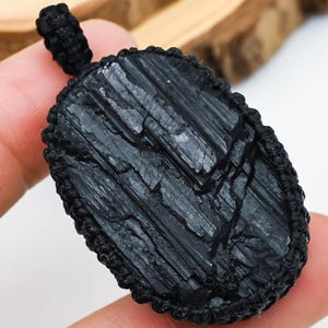 以下が含まれることがあります： 黒トルマリンペンダント、黒い織りコード付き。 生の長方形の石は、マクラメスタイルの織りで囲まれています。 ペンダントは手で持たれています。 テキスト「MAYANK JEWELRY」は左上にあります。