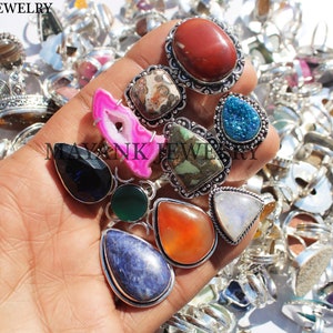 Op de afbeelding: Een verzameling zilveren ringen met verschillende edelsteen bezettingen. De stenen omvatten carneol, amethyst, agaat, turquoise en meer. De ringen worden getoond op een hand.
