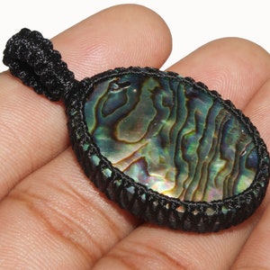 Puede incluir: Un colgante de concha de abulón verde, azul e iridiscente con un cordón negro.