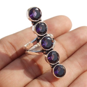 Puede incluir: Un anillo de plata con cinco piedras preciosas púrpuras engastadas en fila. El anillo está diseñado para llevarse en el dedo y tiene una banda larga y fina.