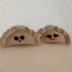May include: Two crocheted brown dumplings with embroidered faces. One dumpling has black eyes and a smiling mouth. The other dumpling has black eyes and a red mouth.