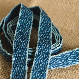 Könnte beinhalten: Nahaufnahme eines blau-weißen gewebten Bandes mit sich wiederholendem geometrischen Muster. Das Band ist auf einer strukturierten, beigefarbenen Oberfläche aufgerollt. Das Bandmuster zeigt miteinander verbundene, stilisierte Formen.