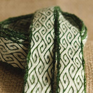 Könnte beinhalten: Ein gewebtes Band in Grün und Weiß mit einem geometrischen Muster. Das Band ist gefaltet und aufgerollt, wodurch das komplizierte Design sichtbar wird.