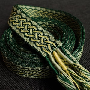 Può includere: Una cintura tessuta nei toni del verde e del giallo, con un motivo geometrico dettagliato. La cintura è arrotolata, con la sezione a fantasia visibile, insieme alle estremità sfilacciate. Il design è intricato e i colori sono naturali.