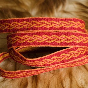 Può includere: Nastro intrecciato rosso e arancione arrotolato, con un motivo a treccia ripetuto. Il nastro è realizzato in un materiale strutturato e viene mostrato su uno sfondo sfocato marrone chiaro. Il nastro è probabilmente utilizzato per scopi decorativi.