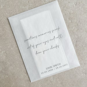 Puede incluir: Un sobre de papel translúcido blanco con el texto "sometimes memories sneak out of your eyes and roll down your cheeks." Debajo, "JOHN SMITH 23.05.1939 - 29.10.2022" en negro. Contiene una tarjeta rectangular blanca.