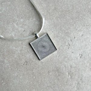 May include: A silver square pendant with a circular design hangs from a white ribbon.