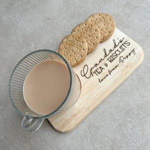 May include: A wooden serving tray with the text "Grandad's Tea & Biscuits Love from Poppy" engraved on it. A glass cup of tea and four biscuits are on the tray.