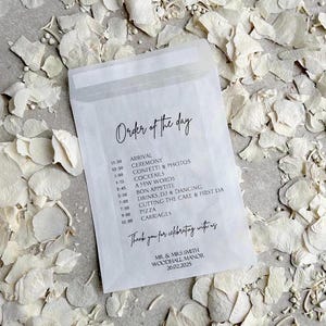 May include: A white paper envelope with a black and white order of the day schedule for a wedding. The schedule includes the arrival time, ceremony, confetti and photos, cocktails, a few words, bon appetite, drinks, DJ and dancing, cutting the cake and first dance, pizza, carriages, and a thank you message for celebrating with the couple. The text reads: "Order of the day", "Arrival 11:30", "Ceremony 12:30", "Confetti & Photos 1:00", "Cocktails 1:15", "A Few Words 2:45", "Bon Appetite 3:30", "Drinks, DJ & Dancing 7:00", "Cutting the Cake & First Dance 7:30", "Pizza 9:00", "Carriages 12:00", "Thank you for celebrating with us", "Mr. & Mrs. Smith", "Woodhall Manor", "26.02.2025".