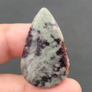 Peut inclure: Une pierre précieuse en forme de goutte d'eau polie. La pierre présente un mélange de motifs vert pâle, violet foncé et noir. La pierre est tenue entre les doigts, mettant en valeur sa taille et sa forme.