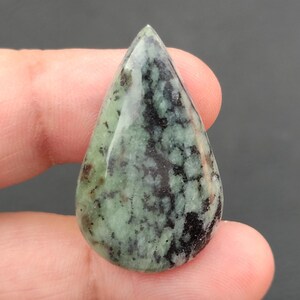 Könnte beinhalten: Ein polierter, tropfenförmiger Edelstein. Der Stein hat ein gesprenkeltes Muster aus Grün und Schwarz. Der Edelstein wird zwischen den Fingern gehalten, was seine Größe und Form zeigt. Die Oberfläche des Edelsteins ist glänzend.