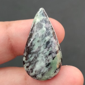 Op de afbeelding: Een druppelvormige edelsteen met een gepolijst oppervlak. De steen heeft een mix van groene, zwarte en witte patronen. De edelsteen wordt tussen vingers gehouden, met een donkere achtergrond.