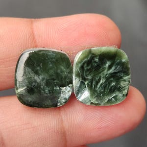 May include: Two square-cut, polished gemstones with a dark green, swirling pattern. The stones are held between fingers, showcasing their translucent quality and natural variations. These gemstones are ideal for jewelry making.