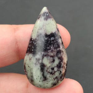 Könnte beinhalten: Ein tropfenförmiger Edelstein mit polierter Oberfläche. Der Stein weist eine Mischung aus hellgrünen, schwarzen und violetten Farbtönen auf, wodurch ein gesprenkeltes Muster entsteht. Der Edelstein wird zwischen den Fingern gehalten und zeigt seine Größe und Form.