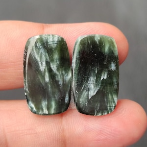 May include: Two polished seraphinite gemstones. Each stone is a dark green color with white, feathery inclusions. The stones have a rectangular shape with rounded corners. The background is a neutral gray.