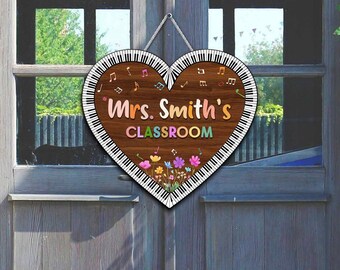 May include: A heart-shaped wooden sign with a piano key border and the text "Mrs. Smith's Classroom." The sign features musical notes and colorful flowers. It hangs on a gray door.
