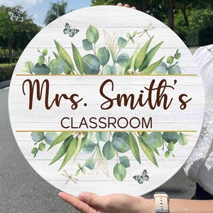 May include: A round, white wooden sign with the text "Mrs. Smith's CLASSROOM" in brown script. The sign is decorated with green eucalyptus leaves, butterflies, and dragonflies. A gold line accents the text.