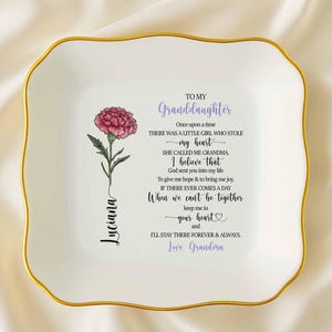 May include: A white ceramic trinket dish with a gold rim and a floral design. The dish features a pink flower and text that reads "To My Granddaughter" and a personalized name. A heartfelt gift.