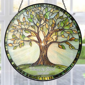 May include: A round stained glass window featuring a tree design. The tree has a brown trunk and branches with green and yellow leaves. The names "Thomas & Sarah" and the date "10.08.2010" are inscribed at the bottom. The window is framed in black and hangs from a chain.