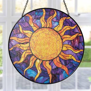 May include: Round stained glass suncatcher with a black metal frame, hanging from a chain. The sun design features a yellow center and orange rays against a background of purple, blue, and teal glass.