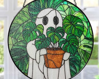 May include: Round stained glass art piece featuring a white ghost holding a potted plant with large green leaves. The background is filled with green leaves. The artwork is framed in a black metal circle and hangs from a chain.
