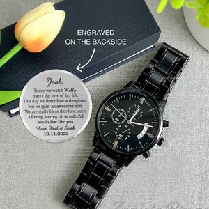 May include: A black watch with a silver engraved message on a round silver disc. The message reads: "Jonh, Today we watch Kelly marry the love of her life. This day we don't lose a daughter, but we gain an awesome son. We are really blessed to have such a loving, caring, & wonderful son-in-law like you. Love, Paul & Sarah 19.11.2022"