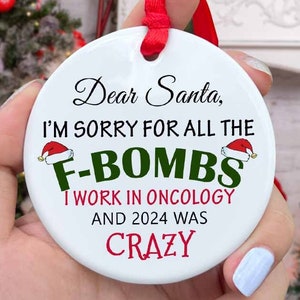 May include: White ceramic Christmas ornament with red ribbon. The ornament features the text "Dear Santa, I'M SORRY FOR ALL THE F-BOMBS I WORK IN ONCOLOGY AND 2024 WAS CRAZY".