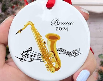 May include: White ceramic Christmas ornament with a gold saxophone, musical notes, and the text "Bruno 2024".