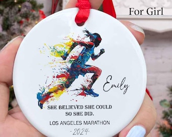 May include: A round white ceramic ornament with a red ribbon hanger. The front features a colorful watercolor-style graphic of a runner, the name Emily, and the text: SHE BELIEVED SHE COULD SO SHE DID. LOS ANGELES MARATHON -2024-.