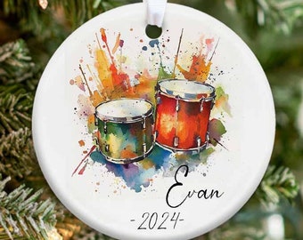May include: A white ceramic Christmas ornament with a watercolor painting of two drums and colorful splatters. The ornament has the name "Evan" and the year "2024" written on it.