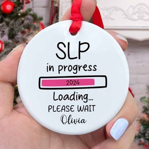 May include: White ceramic Christmas ornament with the text "SLP in progress 2024 Loading... PLEASE WAIT Olivia". The ornament has a pink progress bar and a red ribbon.