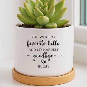 May include: A white ceramic planter with a green succulent plant, sitting on a wooden saucer. The planter has the text "YOU WERE MY favorite hello AND MY HARDEST goodbye" with a paw print and the name "Buddy".