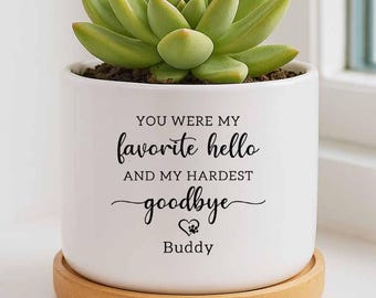 May include: A white ceramic planter with a green succulent plant, sitting on a wooden saucer. The planter has the text "YOU WERE MY favorite hello AND MY HARDEST goodbye" with a paw print and the name "Buddy".