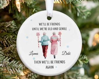May include: White round ceramic ornament with a blue and white snowflake design. The ornament features two women walking with candy canes and the text "We'll be friends until we're old and senile. Then we'll be friends again." The names "Lanna" and "Debi" are written below the women.