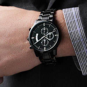 May include: A black chronograph wristwatch with a black leather strap. The watch face has a black background with white markings and a date window.