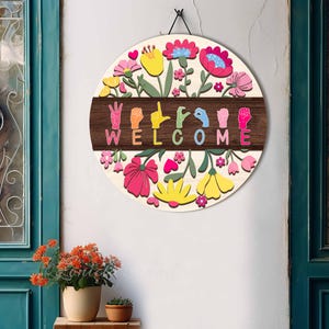 May include: A round wooden welcome sign with a floral design. The sign features colorful flowers in pink, yellow, and blue, surrounding a dark brown band with the word "WELCOME" spelled out in sign language. A black cord is attached for hanging.