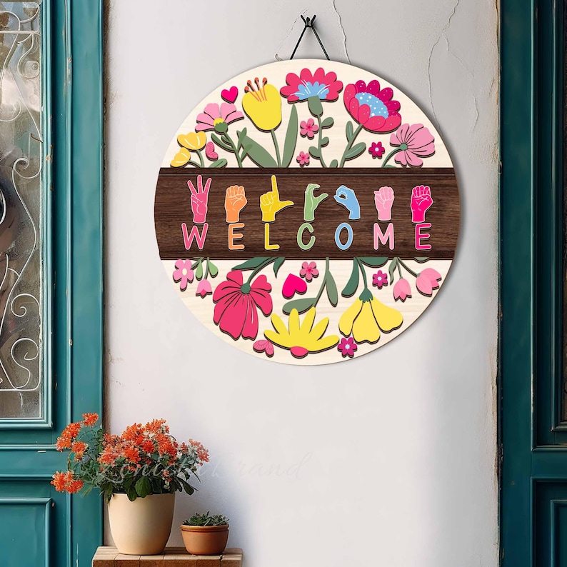 May include: A round wooden welcome sign with a floral design. The sign features colorful flowers in pink, yellow, and blue, surrounding a dark brown band with the word "WELCOME" spelled out in sign language. A black cord is attached for hanging.