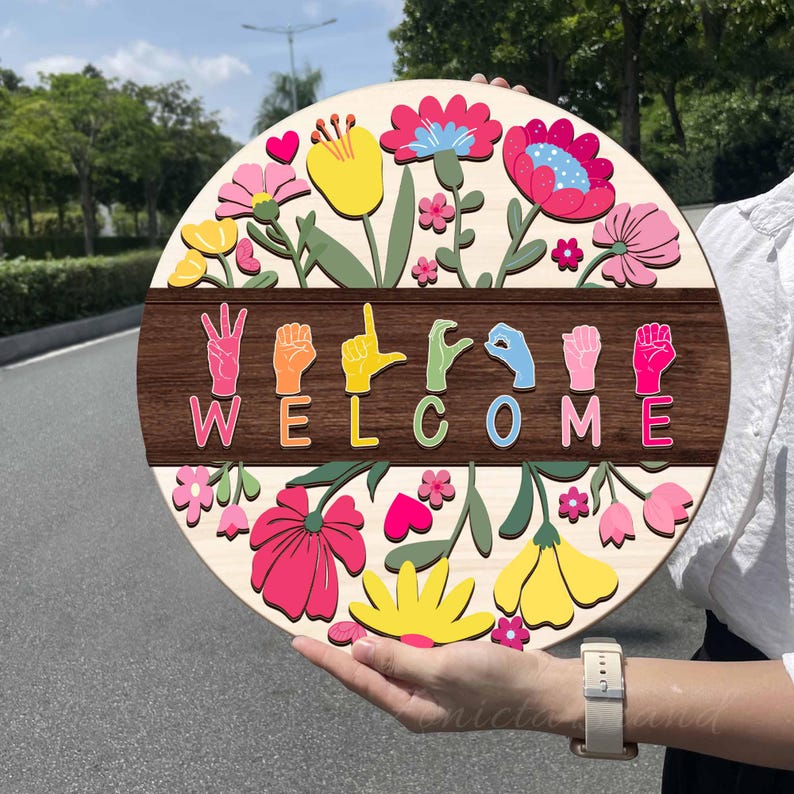 May include: A round wooden sign with a floral design and the word "WELCOME" spelled out in colorful sign language. The sign features pink, yellow, and red flowers with green leaves. The background is a light wood color.