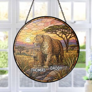 May include: Round stained glass panel featuring a leopard and cub in a savanna landscape. The artwork includes a sunset, trees, and tall grass. The names "THOMAS" and "DADDY" are printed on the bottom. The panel is framed in black and hangs from a chain.