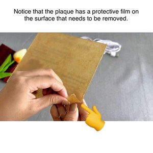May include: A close-up of a hand peeling back a protective film from a brown plaque. The film is being peeled back from the top right corner of the plaque. The text "Notice that the plaque has a protective film on the surface that needs to be removed." is visible above the image.