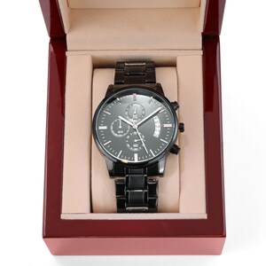 May include: A black chronograph watch with a black leather strap and a date window. The watch is in a red velvet box.
