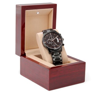 May include: A black chronograph watch with a rose gold face and date window, displayed in a dark red wooden box with a beige interior.