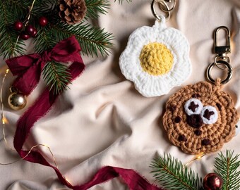 May include: Two crochet keychains featuring a white and yellow fried egg design and a brown chocolate chip cookie design with white eyes and dark brown spots, both attached to gold-toned metal lobster claw clasps and rings.